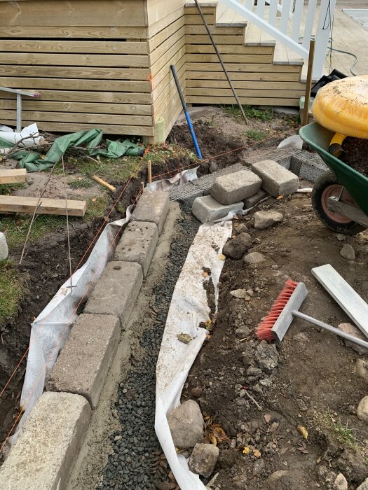 Utspridda murstenar vid en grävd yta med betongsula och grus, bredvid en trappa. Verktyg och material ligger runt arbetsområdet.