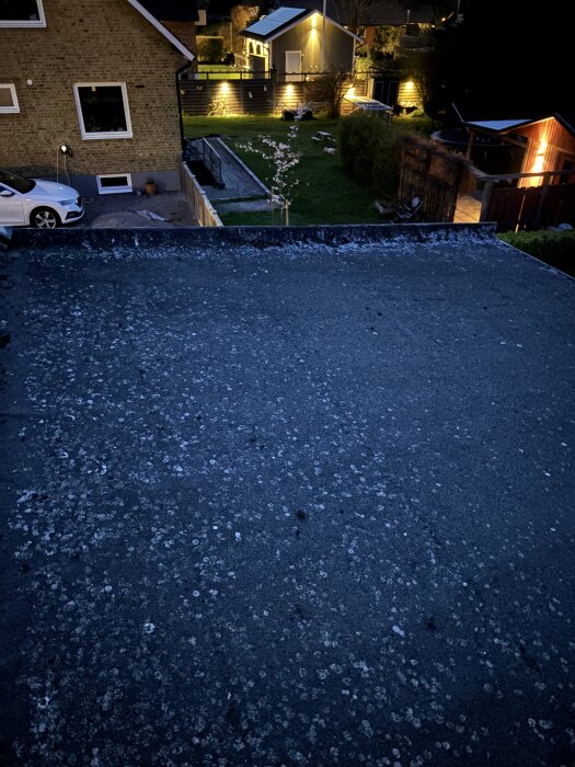 Platt tak fotograferat på kvällen, med asfaltspapp synlig. Omgivande trädgårdar och byggnader är delvis belysta.