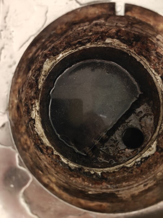 Gammal duschbrunn med delvis rostig kant och liten öppning synlig, antyder avsaknad av normalt vattenlås och svåråtkomligt utlopp.