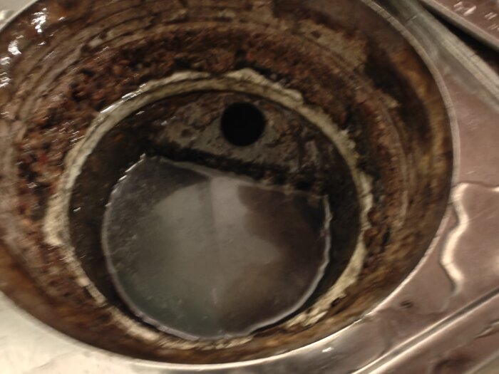 Gammal duschbrunn med avlagringar och smuts, saknar vattenlås. Synlig öppning och hål på sidan. Utlopp till vänster under plastbit.