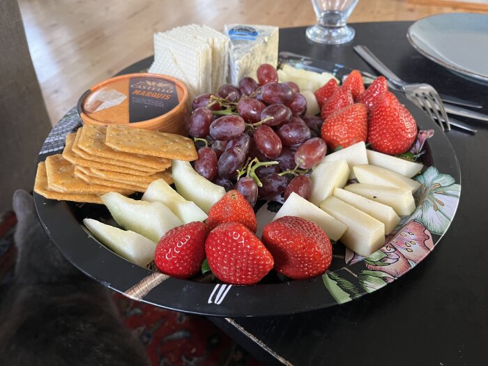 Ost- och fruktbricka med kex, jordgubbar, vindruvor och olika ostar på en blomdekorerad tallrik.