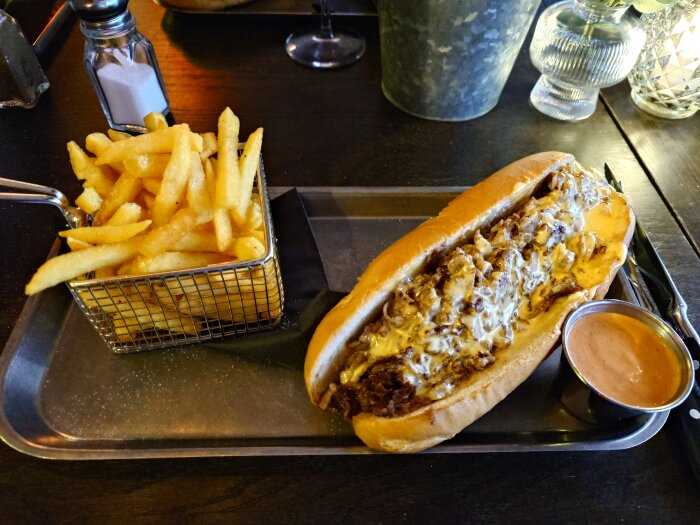 Philly Steak Sandwich med smält ost och kött i bröd serverad med pommes frites i korg och sås på bricka.