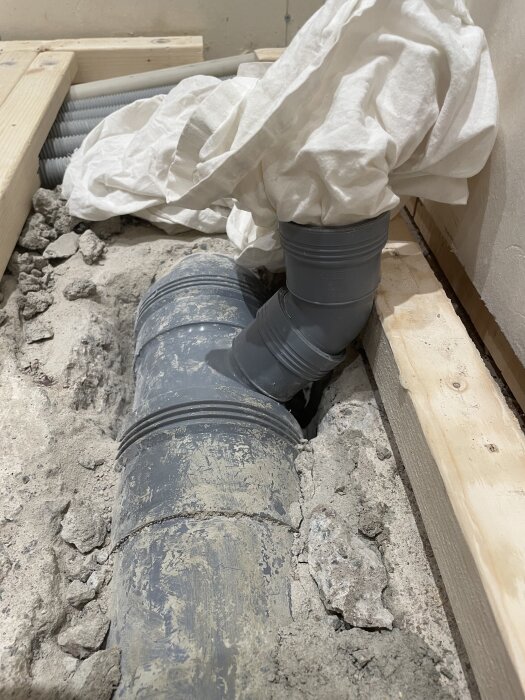 Grå avloppsrörsinstallation med en 45 graders böj och täckt med vit duk, omgiven av betongrester och träreglar.