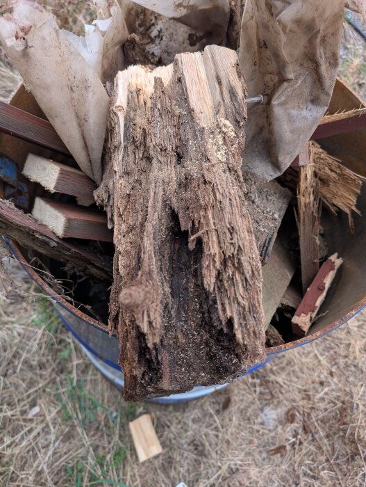 Förmultnad trästolpe i en behållare, omgiven av trasigt papper och träbitar.