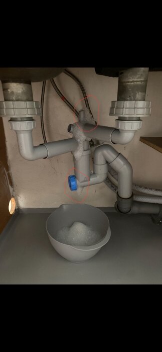 Vattenlås under diskbänk där skum läcker från diskavloppsrör, inringat röda områden visar var läckage sker. Skål placerad under för att samla upp skummet.
