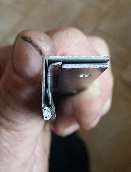 En hand håller i ett modifierat rostfritt gångjärn, med en lång vinkelform, anpassat för användning på nya båtens dörrar.