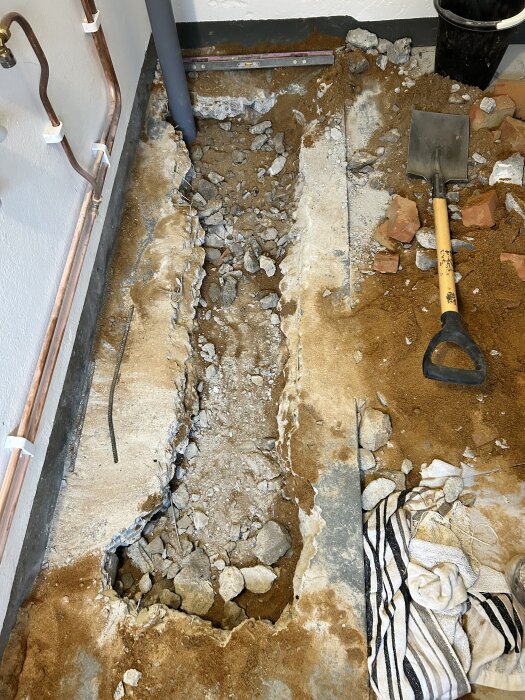 Grävning i betonggolv med synliga rör och stenblock, spade och trasor i bakgrunden, pågående byggprojekt.