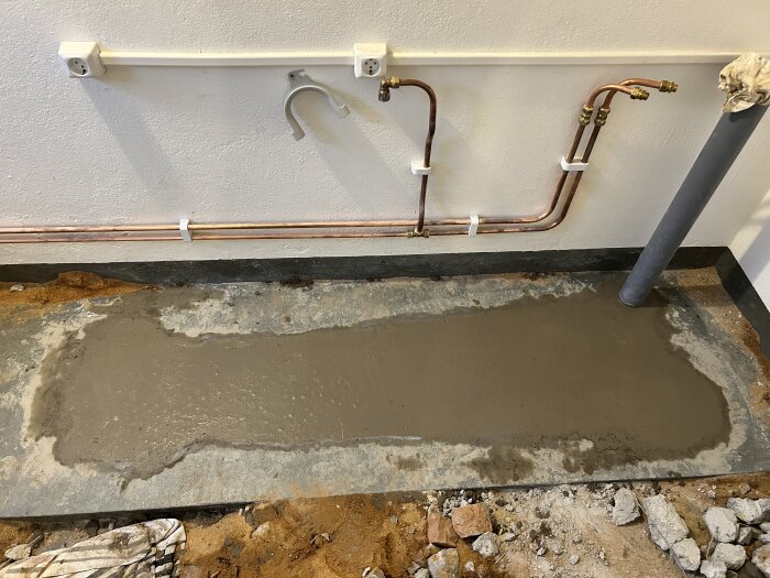 Nyfyllt betonggolv med synliga rörledningar på väggen ovanför och grävda kanter på marken, del av byggprojekt.