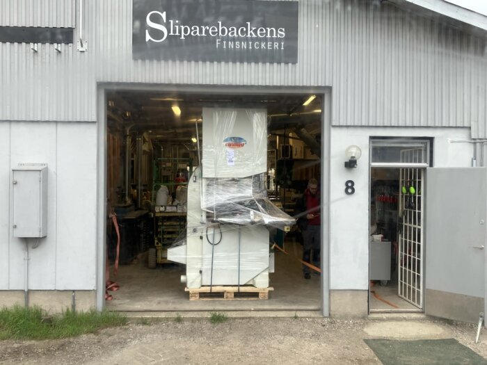 Stor bandsåg inplastad på pall i portar hos Sliparebackens finsnickeri. Placering förberedd för nedlyftning med lastmaskin.