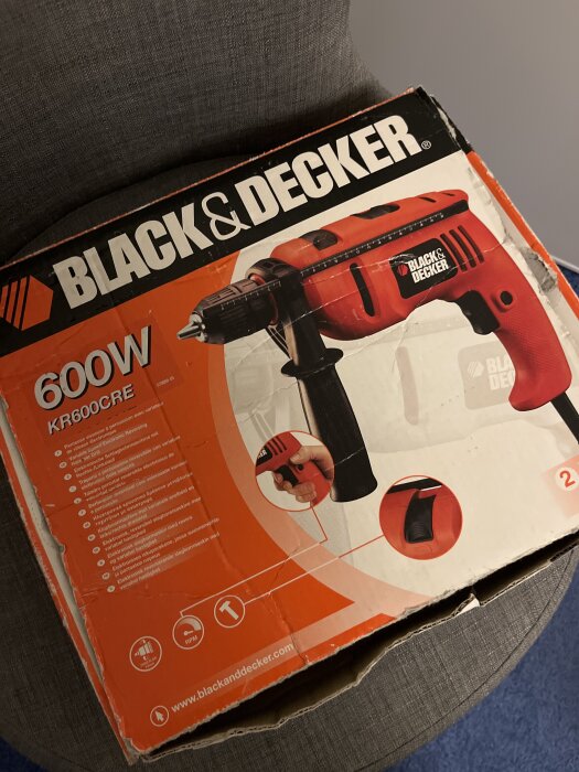 Förpackning till en Black & Decker borrmaskin, modell KR600CRE, 600W, på en grå stol.