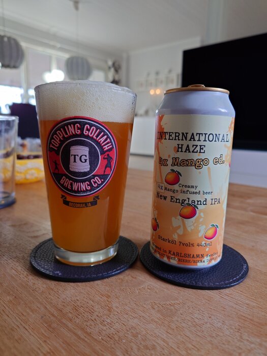 Ölglas med logotyp från Toppling Goliath och en burk "International Haze 4x Mango ed. New England IPA" på ett bord.
