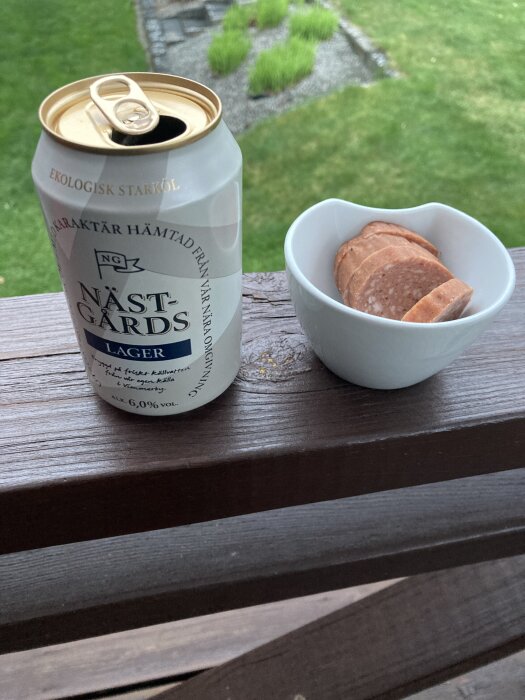 Öppnad burk Nästgårds lager bredvid en skål med skivad korv, placerad på ett trästaket med grön gräsmatta i bakgrunden.