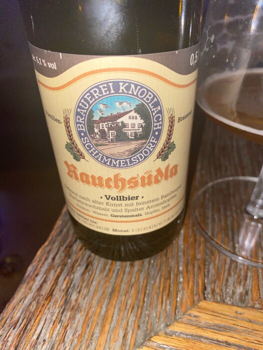 Bilden visar en flaska rököl från Brauerei Knoblach, märkt "Rauchstübl" med ett ölglas bredvid, på ett träbord.