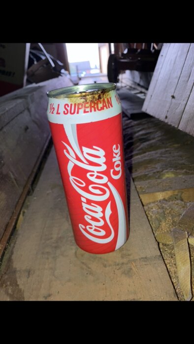 En gammal Coca-Cola Supercan-burk står på ett dammigt golv på en vind. Burken ser ut att vara från en äldre tid.