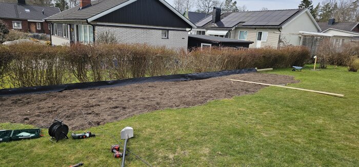 Trädgårdsområde med nyplanerad jord för gräs, omgiven av buskar, verktyg och fuktisoleringsduk.