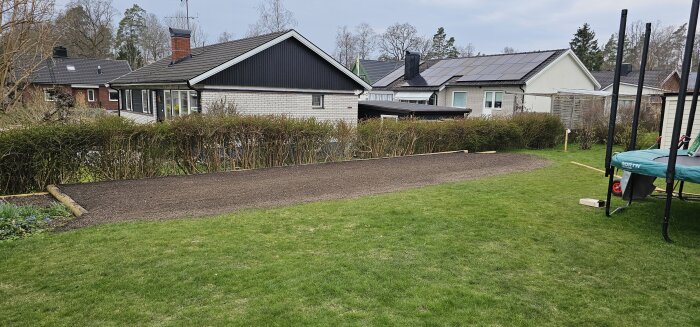 Nyplanerad grusyta med armerade timmerkanter omgiven av gräsmatta, redo för gräsfrösådd med utsikt över villaträdgårdar.