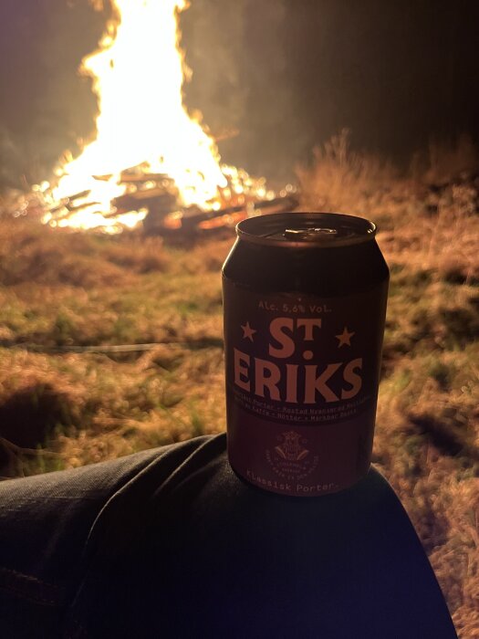 En ölburk med texten "St Erik" framför en stor brasa på gräs, kvällstid.