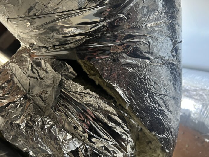Lös stenullsisolering och aluminiumtejp uppdelat kring rör på kallvind, delvis lossnat, synlig isolering utan skydd.