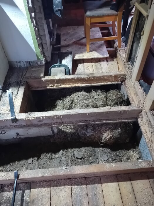 Öppnat golv i stuga med synlig jord och rörigt material under, träreglar och ett trälock intill. En stol står på det delvis exponerade golvet.