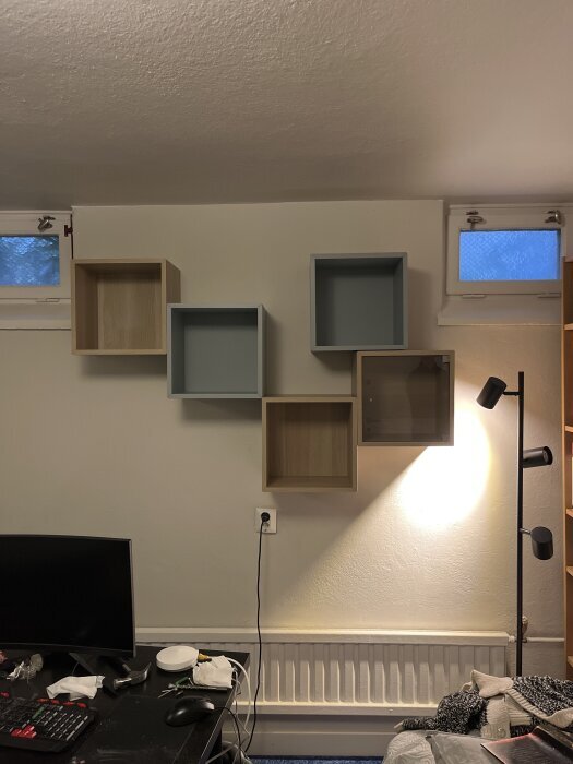 Väggmonterade hyllor i olika färger och storlekar, monterade på vit vägg ovanför skrivbord med dator och lampa.