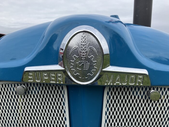 Blå traktor med texten "Super Major" på fronten, bredvid ett märke med vete och en växelsymbol.