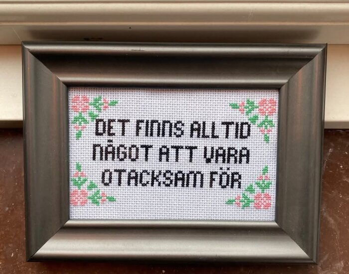 Broderad tavla med texten "Det finns alltid något att vara otacksam för" i en grå ram, dekorerad med blommotiv.