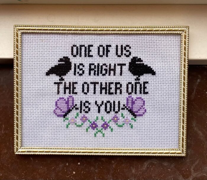 Textbroderi i ram med orden "One of us is right, the other one is you" och motiv av två svarta fåglar samt blommor i lila och grönt.