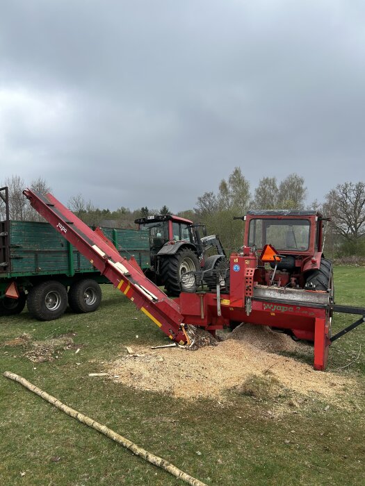 Röd vedklyvningsmaskin används på gräsmatta, omgiven av sågspån och träbitar, med traktor i bakgrunden.
