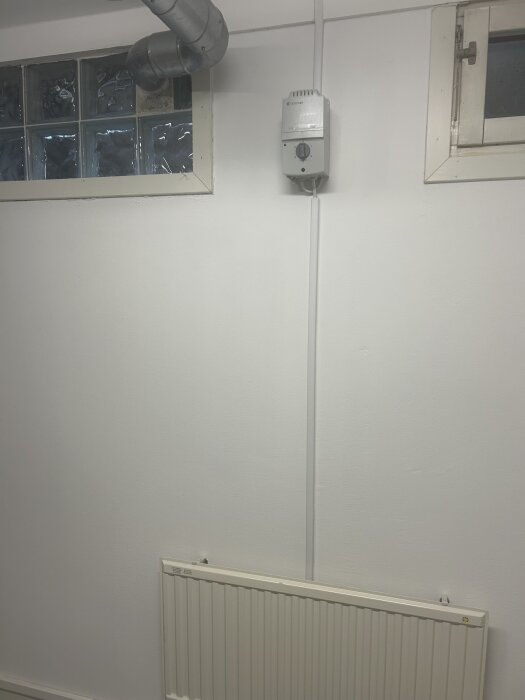 Vitt väggmonterat element och transformator i källare, under ett fönster med vita ramar och ventilationsrör.