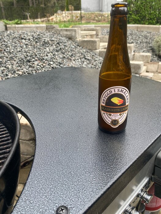 Flaska med lageröl, "Bryggd i Småland Glasrikets Lager", står på en utegrill bredvid en trädgård med grus och stentrappa i bakgrunden.