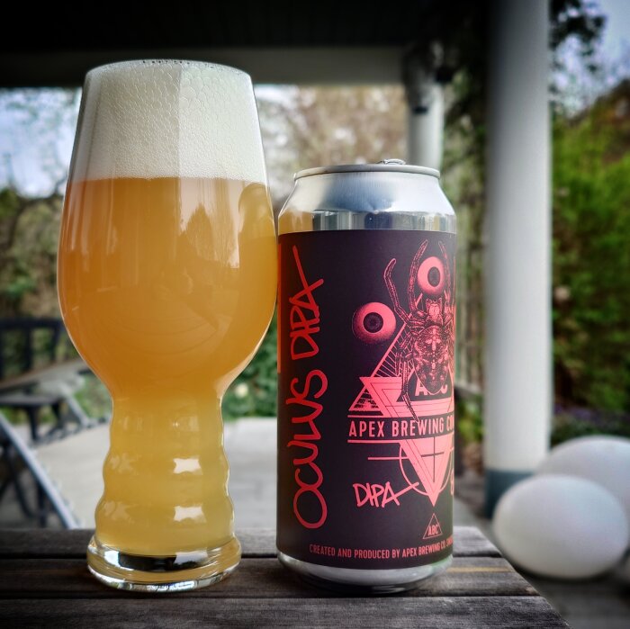 Ett glas och en burk med öl från Apex Brewing Co på ett bord, omgivna av grönska.