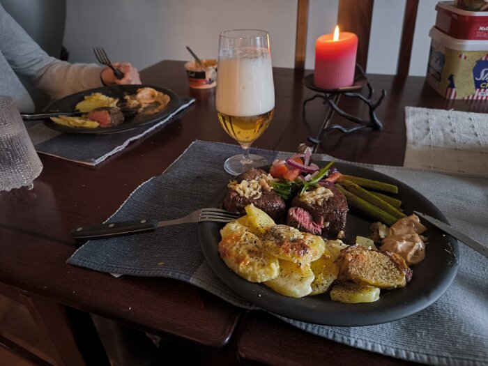 En tallrik med grillad oxfilé, potatisgratäng, smörstekt sparris, pepparsås och sallad. Ett glas öl och en brinnande röd ljus står på bordet.