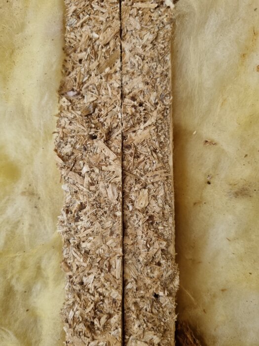 Spånskivsrester sitter fast på en träregel, omgiven av gul isolering.