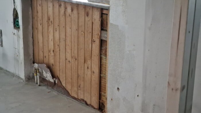 Träpanel i köket vid fönstret, delvis skadad, med rörgenomföring i nederkant och isolering synlig bakom.