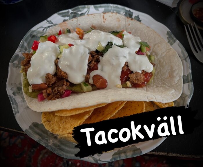 Taco med grönsaker, köttfärs, salsa, vitlökssås, guacamole och picklad rödlök på tortillabröd, serverat ovanpå nachos. Text: "Tacokväll".