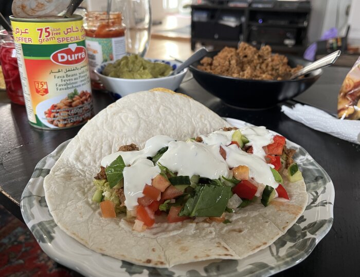 Två tortillabröd fyllda med grönsaker, köttfärs och vitlökssås. Burk med favabönor och skålar med guacamole och salsa i bakgrunden.