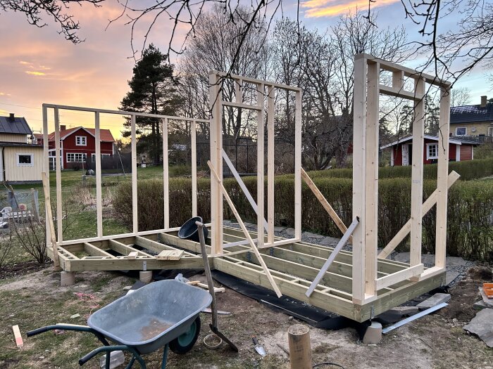 Träramarna för en byggnad är resta på en gård. I förgrunden finns en skottkärra och byggmaterial. Solnedgång i bakgrunden.