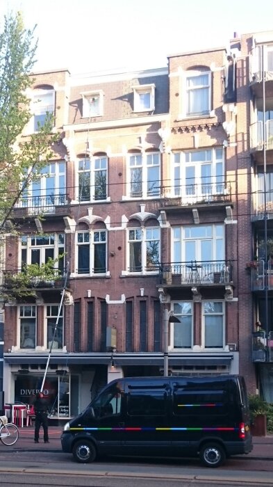 Fönsterputsare med lång stång rengör fönster på flerbostadshus i Amsterdam; svart skåpbil och cykel framför byggnaden.