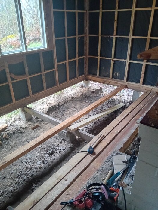 Renovering pågår med synliga bjälkar och verktyg på ett grusgolv. Fönster och väggar utan isolering i ett rum med åsikt om plast.