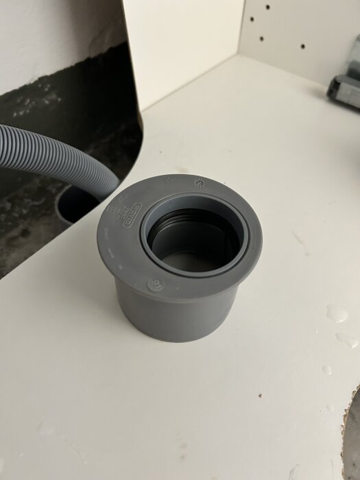 Grå adapter för VVS-rör på vit yta, med ett flexibelt rör synligt i bakgrunden.