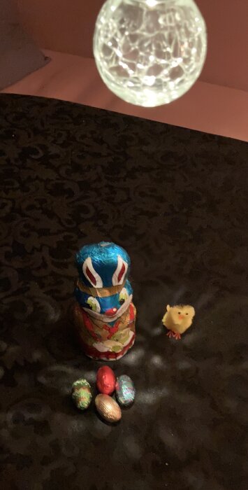 Chokladkanin och chokladägg på ett mörkt bord, med en liten dekorativ kyckling bredvid och ett hängande ljus ovanför.