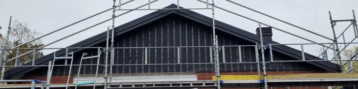 Byggnadsställningar runt ett hus under takrenovering. En ny lockpanel på gavelspetsen ser kort ut, med synliga hål under den.
