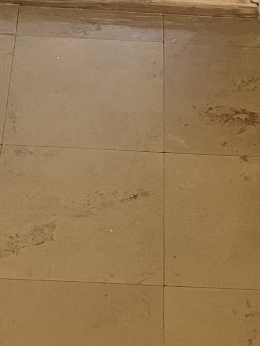 Golv med tunna, hårda plattor, vissa skadade, avslöjar svart material under; misstänks innehålla asbest, original från 1952.