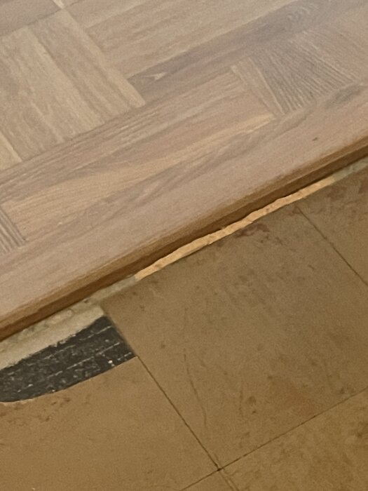 Gammalt golv med trasiga, tunna plattor ovanpå svart underlag, synligt under nylagt laminatgolv i en lägenhet.