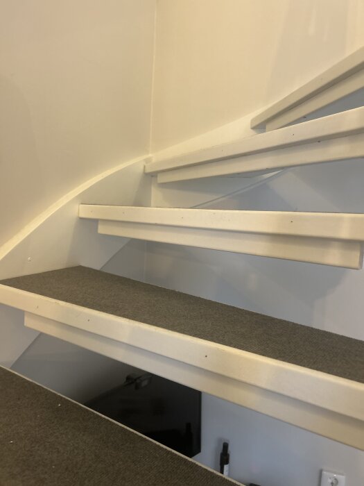 Vitmålad trappa med ny matta på stegen. Undersidan av trappan har en list fäst med skruv. Gulaktigt ljus i rummet.