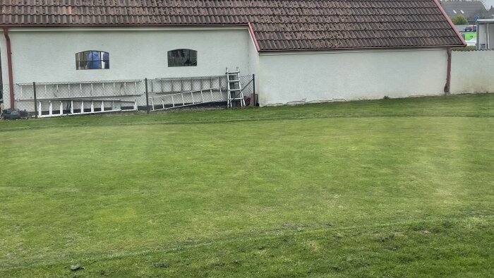 Nyligen klippt gräsmatta framför vit byggnad med takpannor, stege lutad mot staket. Backend av trädgård syns också.