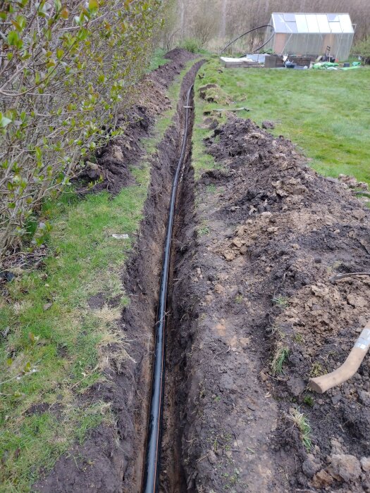 Nedgrävd PEM-slang i grävd dike med grönska till vänster och ett växthus i bakgrunden. Jorden är uppgrävd längs diket.