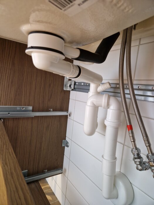 Nedre del av ett handfat med vita avloppsrör i plast anslutna till väggen och flätade metallslangar bredvid i ett badrum.