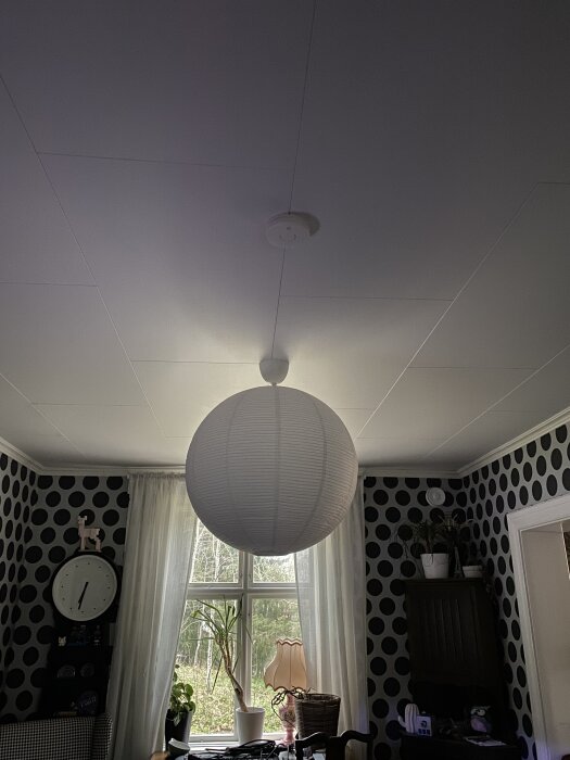 Taklampa och brandvarnare installerade i ett rum med mönstrad tapet och fönster med växter i fönsterkarmen.