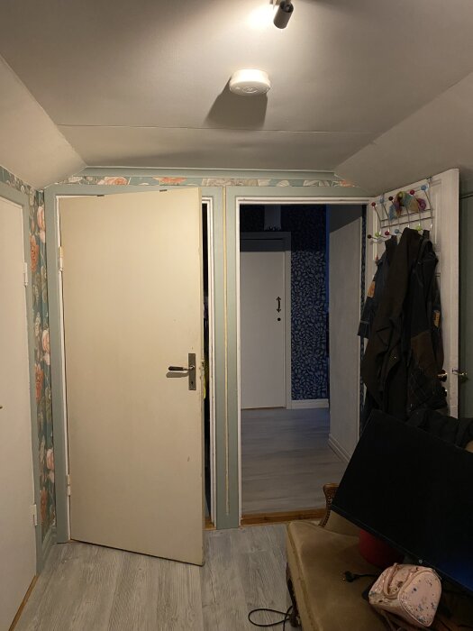 Brandvarnare i taket i en hall med vita dörrar, färgglad klädhängare och mönstrad tapet på väggarna.